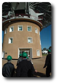 People with green hard hats in front of circular brown building below telescope dish