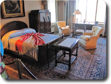 Bedroom with double bed and dress draped over it; a couple of chairs and writing desk