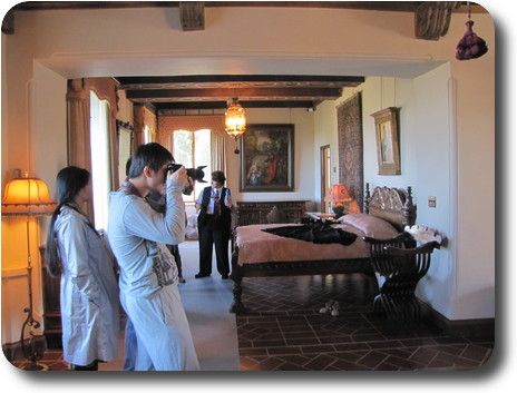 Bed in large room, with photographer; paintings on walls, dress on bed