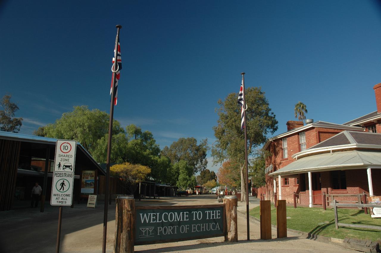 Welcome sign across dirt road with historical buildings and trees behind