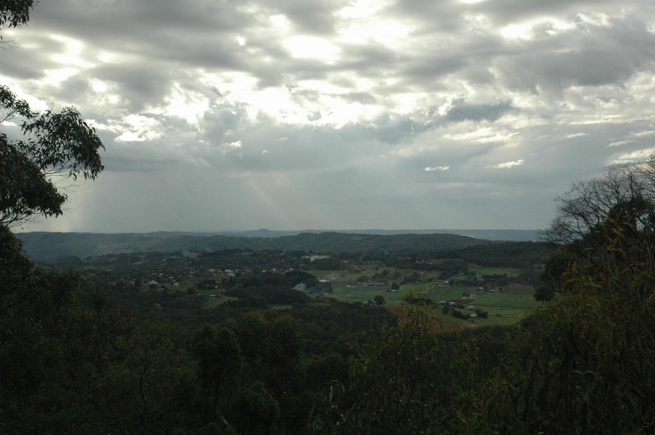 Distant green fields between close trees; grey skies with some rain visible