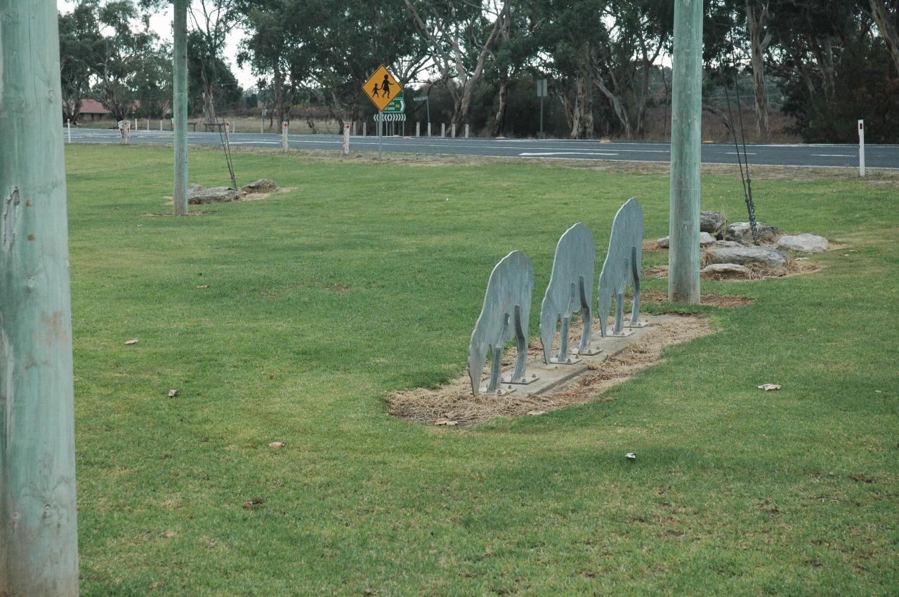Two wooden poles, metal shapes between in grassy area with road in background