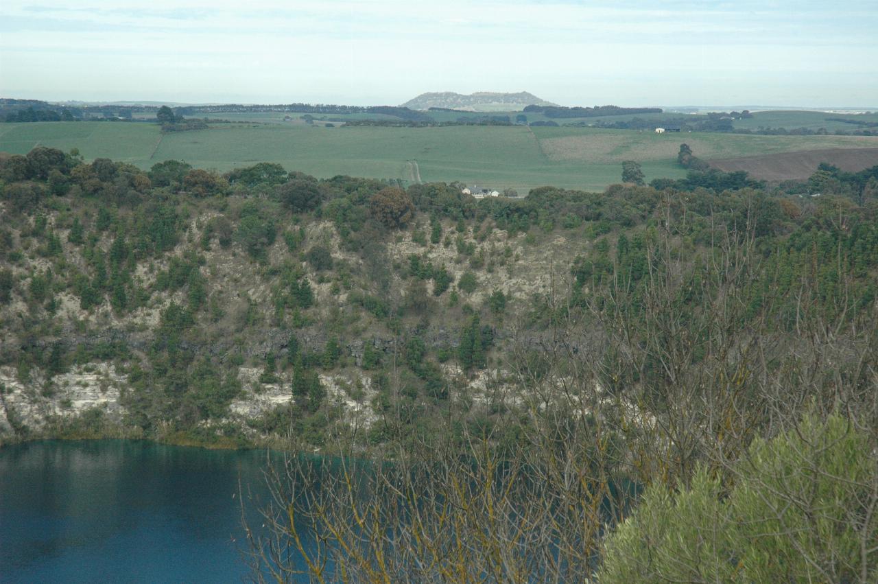 Part of water filled volcanic crater, green fields and hump of mountain in the distance