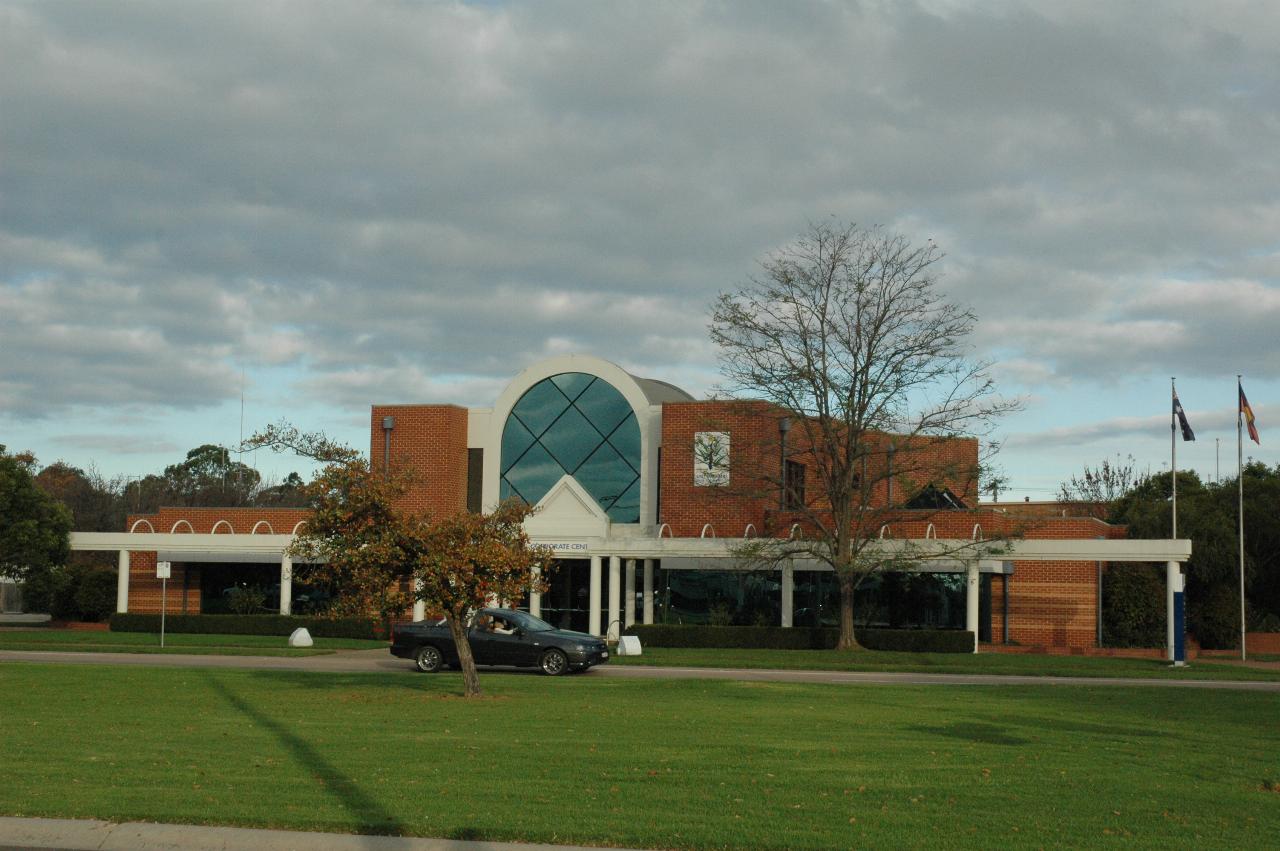 Modern building with arched central roof and wings, seen across wide median strip
