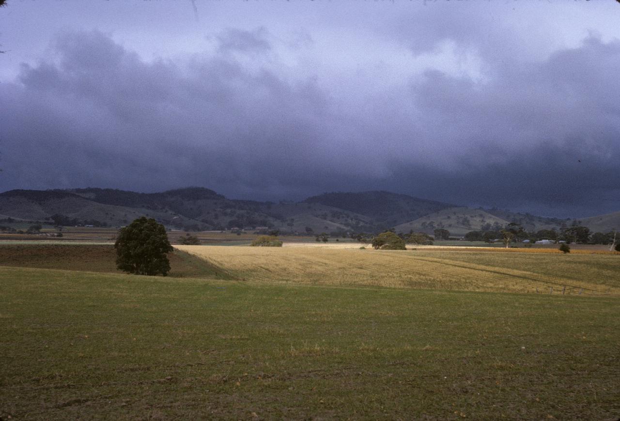 Sun shining on close, brown fields, distant hills in shadow and under dark clouds